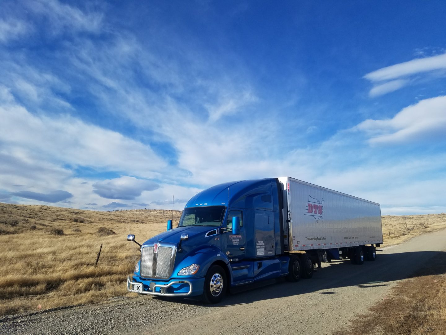 Picture of the left side of a blue Kenworth Semi-truck pulling a DTS reefer trailer on a dirt road, in the country, with blue skies above.